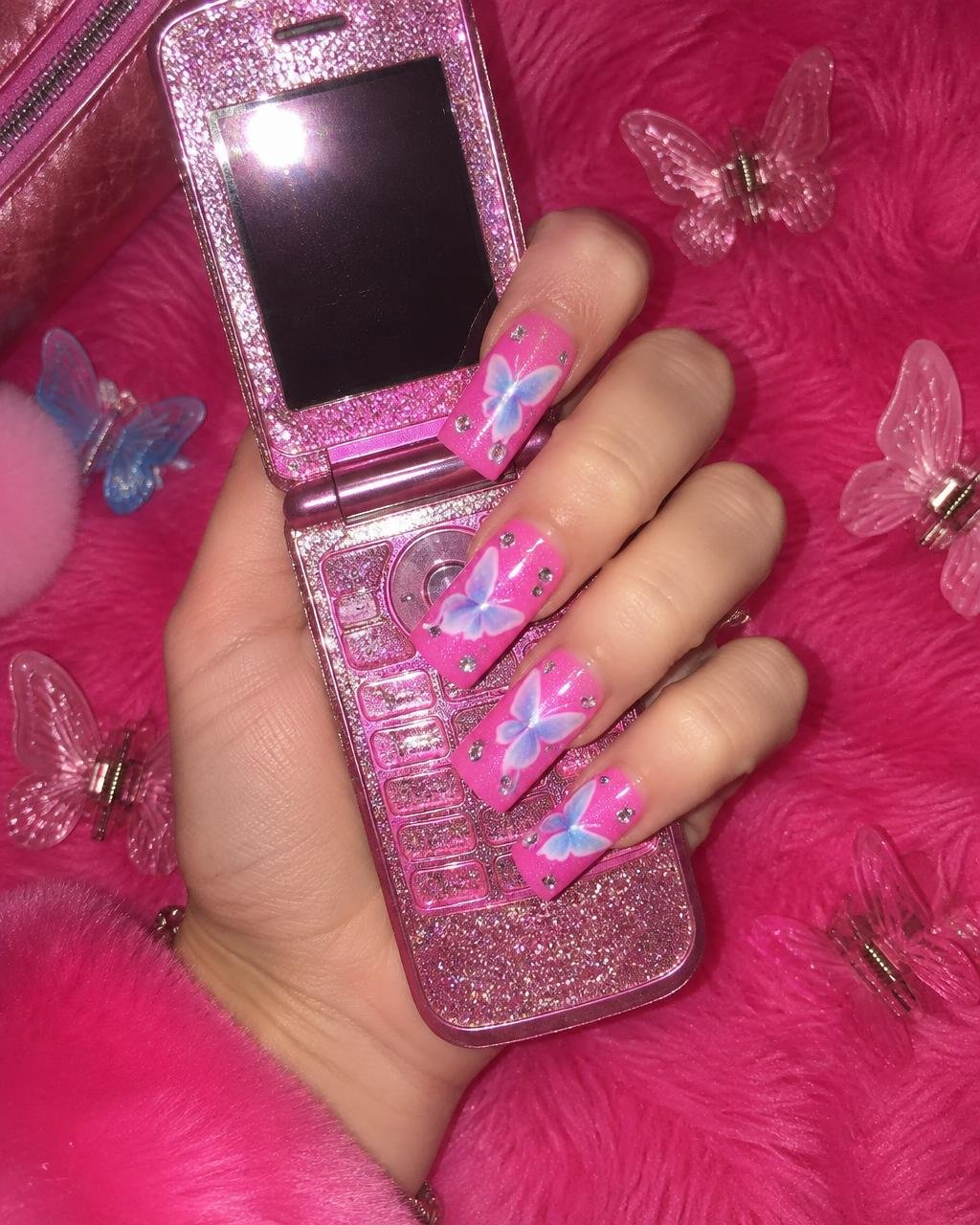 Hot pink square nails with airbrushed blue butterflies and rhinestones holding a glitter flip phone
