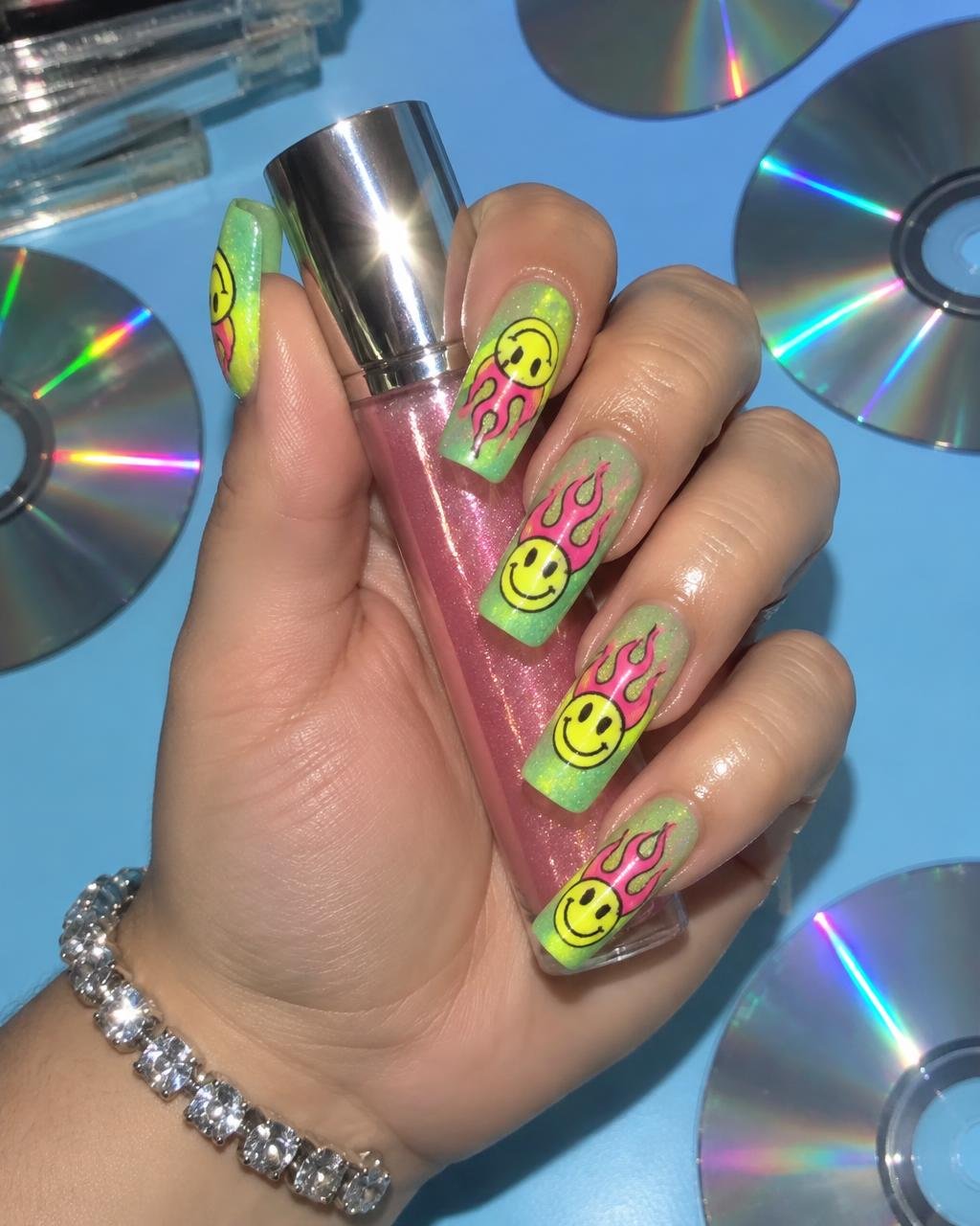 Lime coffin nails with airbrushed smiley faces and pink flames holding lip gloss over CDs